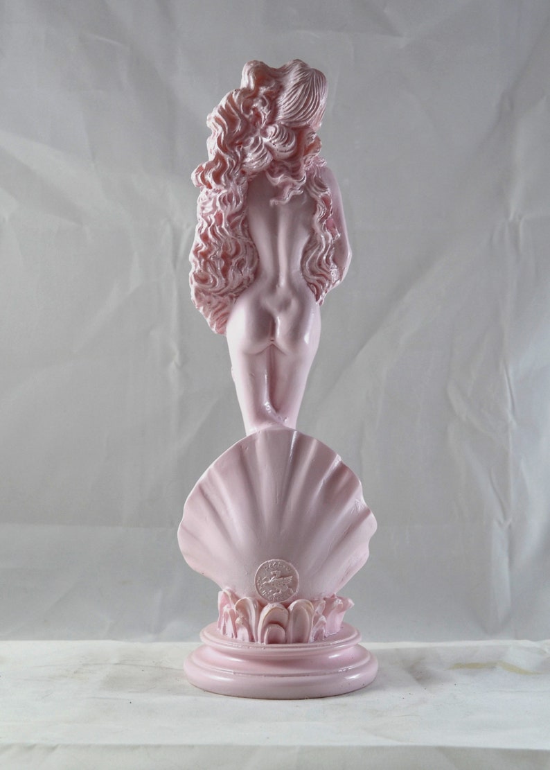 May include: A pink ceramic statue of the Venus de Milo standing on a shell base. The statue is a replica of the famous Greek sculpture.
