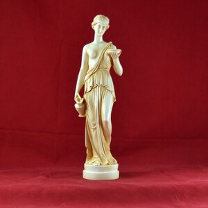 May include: A cream-coloured statue of a classical female figure, holding a small bowl and a jug. The figure is draped in flowing robes and stands on a round base. The background is a solid red.