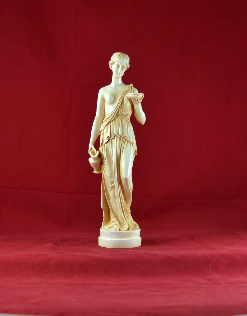 May include: A cream-coloured statue of a classical female figure, holding a small bowl and a jug. The figure is draped in flowing robes and stands on a round base. The background is a solid red.