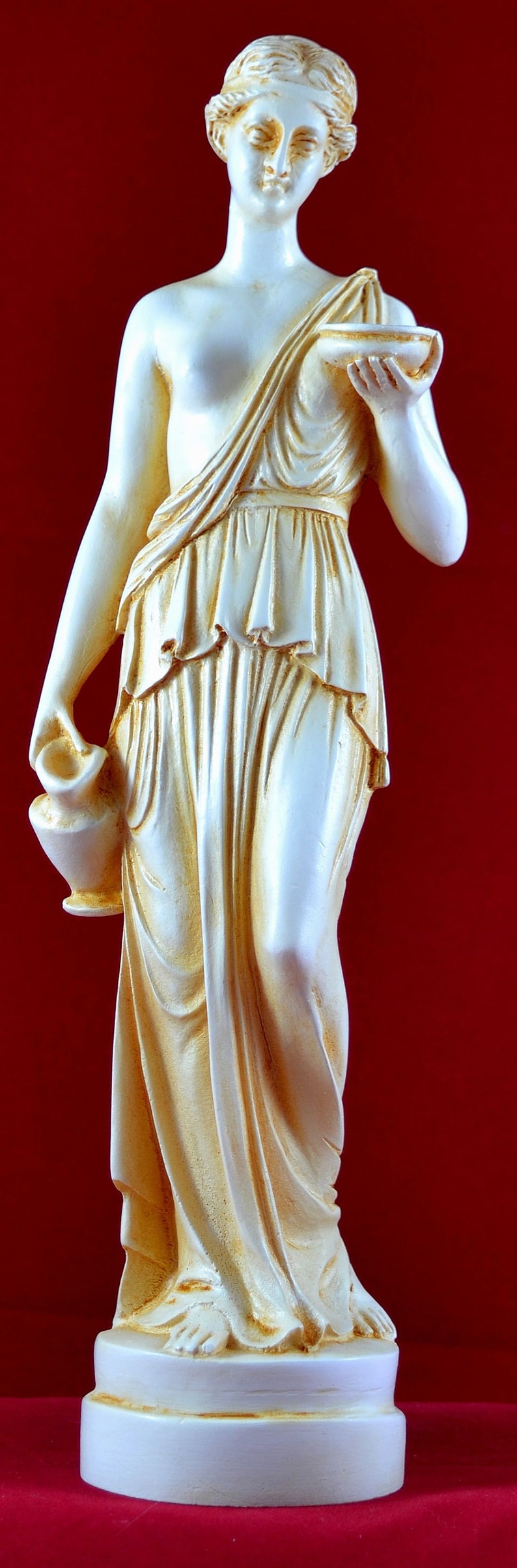 May include: A cream-coloured statue of a classical female figure, holding a small bowl and a vase. The statue is draped in flowing robes and stands on a white base against a red background.