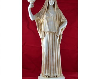May include: A cream-colored statue of a woman in classical attire, holding a chalice aloft. The figure is draped in flowing robes and a head covering. The base of the statue has the Greek word "ESTIA" inscribed.