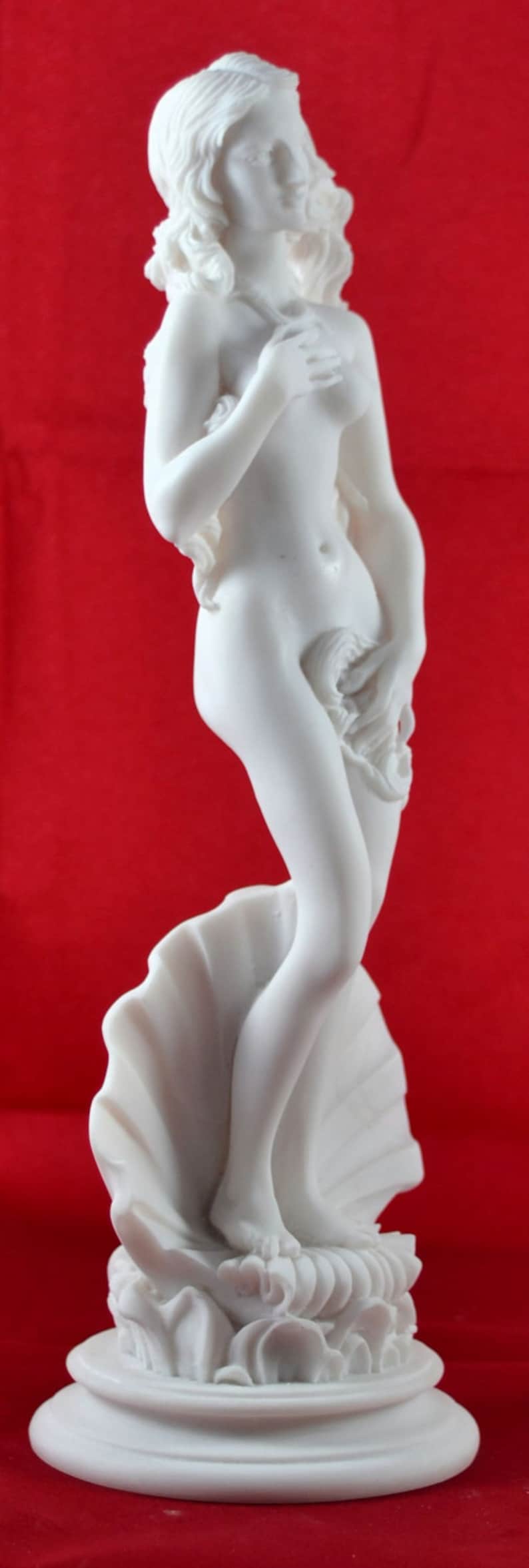 May include: White marble statue of a woman standing on a seashell. The woman is nude and has long flowing hair. The statue is on a white marble base.