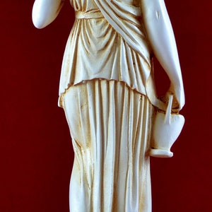 May include: A cream-coloured statue of a classical figure, viewed from the back, holding a small jug. The figure is draped in flowing robes. The statue is on a base with a circular emblem. The background is a deep red.