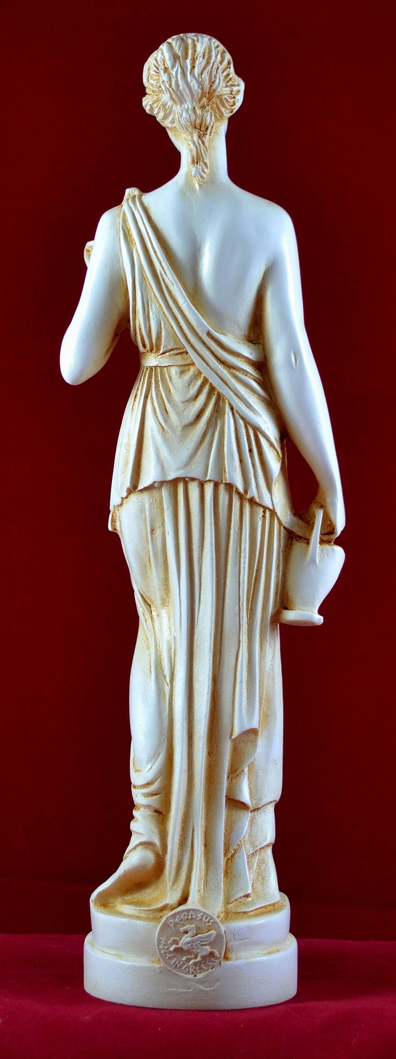 May include: A cream-coloured statue of a classical figure, viewed from the back, holding a small jug. The figure is draped in flowing robes. The statue is on a base with a circular emblem. The background is a deep red.