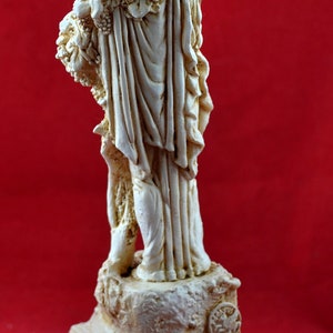 Op de afbeelding: Cr&egrave;mekleurig beeld van een klassieke figuur die een beker omhoog houdt. De figuur is versierd met wijnranken en gehuld in vloeiende gewaden. Het beeld staat op een vierkante basis met een Grieks sleutelpatroon.
