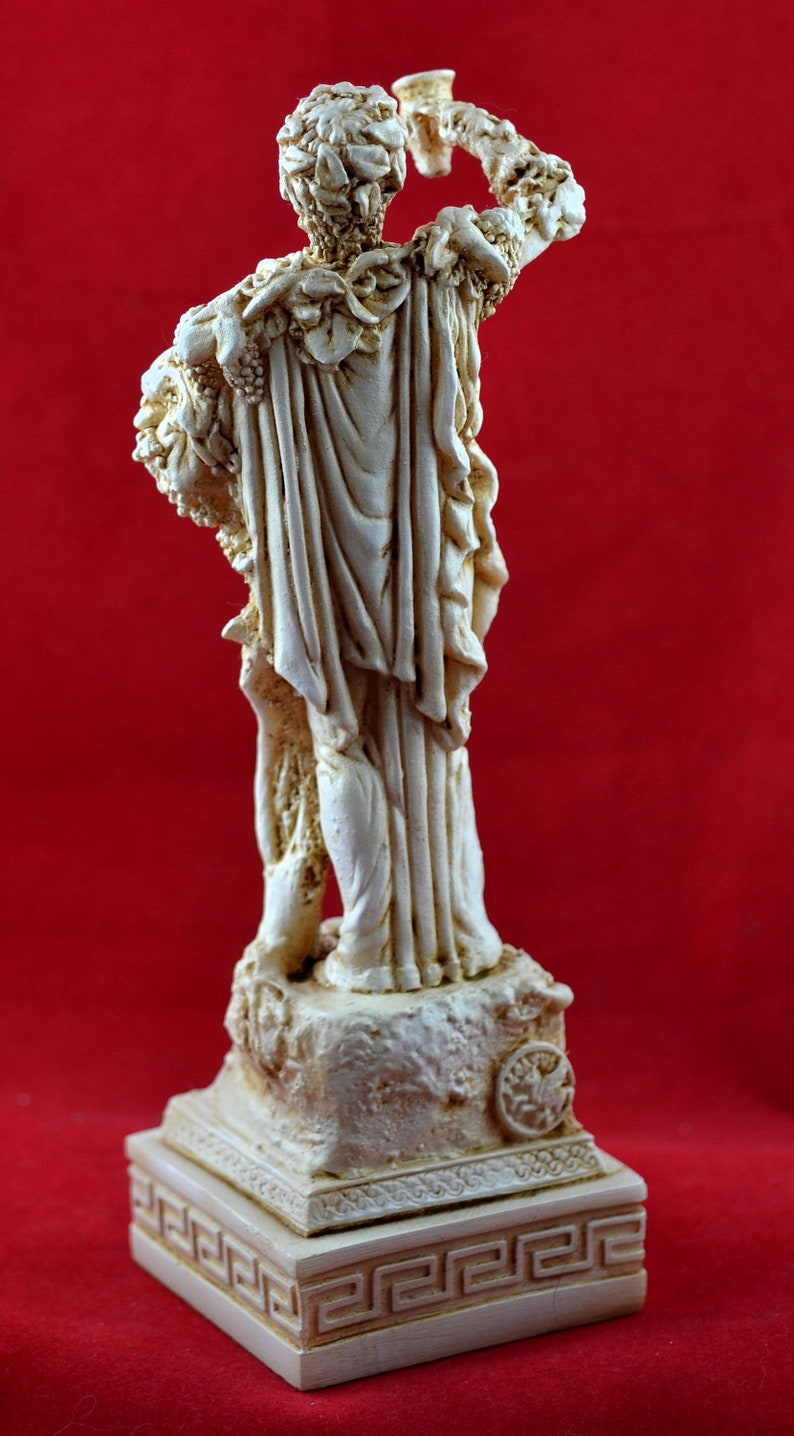 Op de afbeelding: Cr&egrave;mekleurig beeld van een klassieke figuur die een beker omhoog houdt. De figuur is versierd met wijnranken en gehuld in vloeiende gewaden. Het beeld staat op een vierkante basis met een Grieks sleutelpatroon.
