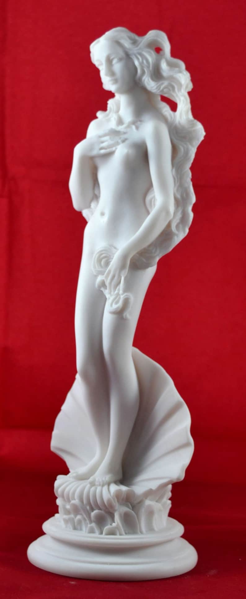 May include: White marble statue of a woman standing on a shell. The woman is nude and has long flowing hair. The statue is on a white base.