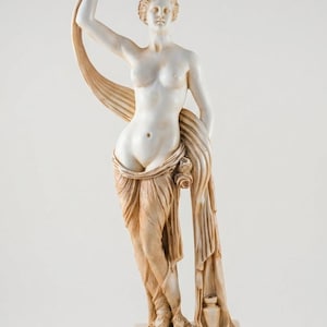 Könnte beinhalten: Eine weiß-beige marmorierte Statue einer stehenden Figur mit erhobenem Arm, die im unteren Körperbereich mit Stoff drapiert ist und auf einem rechteckigen Sockel mit der griechischen Inschrift ΑΦΡΟΔΙΤΗ an der Vorderseite steht.