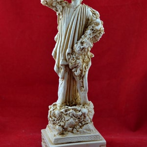 Op de afbeelding: Cr&egrave;mekleurig beeld van Dionysus, de Griekse god van de wijn, op een rode achtergrond. Het beeld houdt een beker vast en is versierd met wijnranken. De basis van het beeld is gegraveerd met de naam "DIONYSUS".