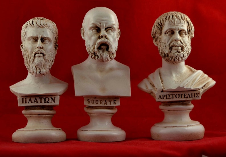 May include: Three white plaster busts of famous Greek philosophers: Plato, Socrates, and Aristotle. Each bust is on a white pedestal with the philosopher's name inscribed in Greek.