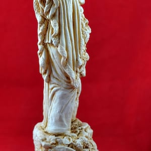 Op de afbeelding: Cr&egrave;mekleurig beeld van een klassieke figuur met een hoorn, tegen een rode achtergrond. Het beeld heeft gedetailleerde draperie&euml;n en is gemonteerd op een decoratieve basis met een Grieks sleutelontwerp.