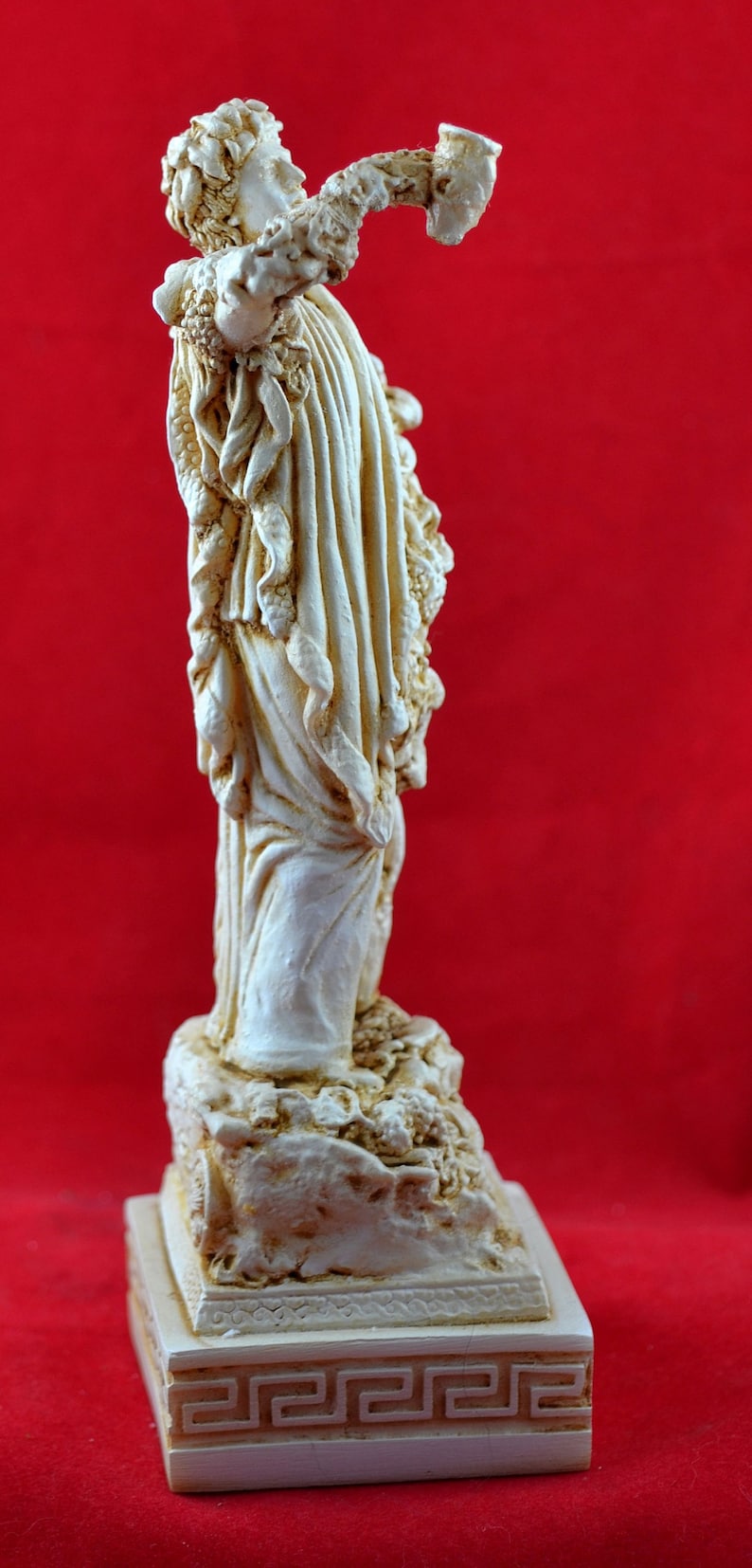 Op de afbeelding: Cr&egrave;mekleurig beeld van een klassieke figuur met een hoorn, tegen een rode achtergrond. Het beeld heeft gedetailleerde draperie&euml;n en is gemonteerd op een decoratieve basis met een Grieks sleutelontwerp.