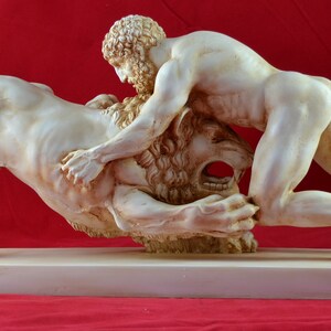 以下が含まれることがあります： 男性がライオンと格闘している白い大理石の彫刻。男性はライオンの上にいて、ライオンの頭を抑えている。彫刻は白い大理石の台座の上に置かれている。