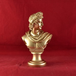Könnte beinhalten: Eine goldene Büste eines Mannes mit einem Lorbeerkranz auf dem Kopf. Die Büste steht auf einem goldenen Sockel mit der Inschrift "APOLLON" auf der Vorderseite.