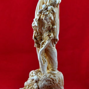Op de afbeelding: Ivoor kleurig beeld van een figuur die een beker vasthoudt, mogelijk een afbeelding van een Griekse god. De figuur is gehuld in vloeiende gewaden en staat op een decoratieve basis met een Grieks sleutelpatroon. De achtergrond is effen rood.