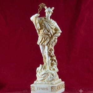 Peut inclure: Une statue blanche et dorée représentant une silhouette portant un casque ailé et des sandales, tenant un caducée. La silhouette est drapée dans une tunique et une cape, debout sur une base rectangulaire avec le texte grec "ΕΡΜΗΣ" et une bordure géométrique.