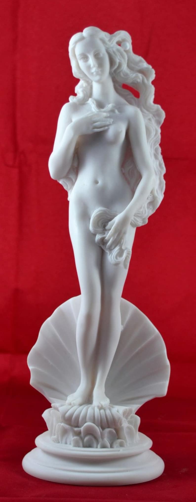 May include: White marble statue of a woman standing on a seashell. The woman is nude and has long flowing hair. The statue is on a white marble base.