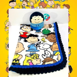Puede incluir: Una manta blanca con un borde de ganchillo azul y negro presenta un estampado de personajes de Peanuts. La manta se acompaña de un personaje de peluche de Charlie Brown.