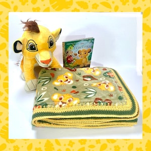 May include: A plush Simba toy, a copy of "The Lion King" book, and a green blanket with Simba and leaf patterns. The blanket has a yellow crocheted border and green trim. The items are arranged on a white surface.