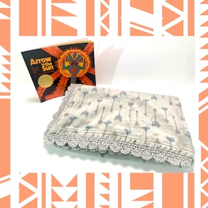 May include: A gray and white baby blanket with a crocheted border and a pattern of arrows. The blanket is next to a book titled "Arrow to the Sun" with a colorful illustration on the cover.