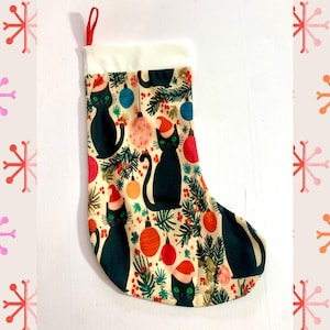 May include: Christmas stocking with a cream background featuring black cats wearing Santa hats, ornaments, and greenery. The stocking has a red loop for hanging and a white cuff.