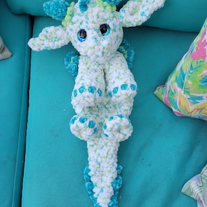 Puede incluir: Un juguete de dragón de peluche hecho a mano en blanco, turquesa y verde. El dragón tiene ojos grandes y redondos, una cola larga y alas pequeñas. El peluche está hecho de tela suave y texturizada y está sobre una superficie turquesa.