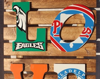 May include: Wooden sign spelling "LOVE" with each letter decorated with a different Philadelphia sports team logo. The "L" is green with the Philadelphia Eagles logo, the "O" is blue with the Philadelphia Phillies logo, the "V" is orange with the Philadelphia Flyers logo, and the "E" is white with the Philadelphia 76ers logo.