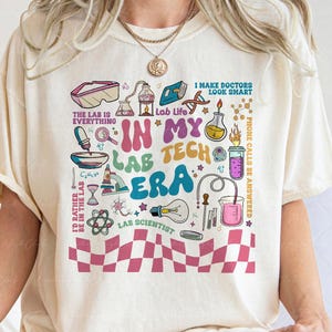 May include: Cream-colored t-shirt with a colorful graphic design featuring lab-related illustrations and text. The text includes "IN MY LAB TECH ERA" and other phrases like "Lab Life" and "Lab Scientist". A pink and white checkered pattern is at the bottom.