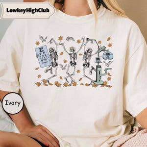 Könnte beinhalten: Ein elfenbeinfarbenes T-Shirt mit einem Grafikdruck von drei tanzenden Skeletten zwischen herbstlichen Blättern und Fledermäusen. Ein Skelett hält eine Sehtafel, während ein anderes mit einem Optometriegerät interagiert. Der Text LowkeyHighClub steht oben.