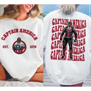 May include: White sweatshirt featuring the text "CAPTAIN AMERICA" in red arched lettering. The front design includes a circular graphic with a character illustration and the text "EST. 1978". The back of the sweatshirt repeats "CAPTAIN AMERICA" with a character illustration.