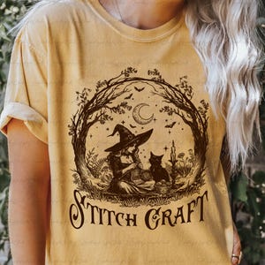 May include: A mustard yellow t-shirt featuring a dark brown graphic print of a witch wearing a pointed hat and knitting, accompanied by a black cat, a crescent moon, bats, and trees. The text STITCH CRAFT is printed in a decorative font below.