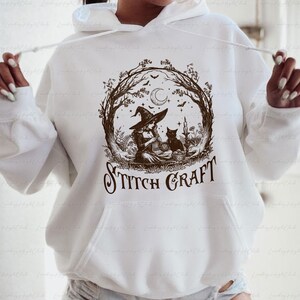 May include: A white hooded sweatshirt featuring a brown graphic print of a person in a witch hat knitting, accompanied by a black cat, a crescent moon, and trees. The text STITCH CRAFT is printed in a decorative font below the illustration.