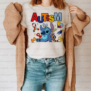 Op de afbeelding: Een wit T-shirt met het woord "AUTISME" in regenboogkleuren en een blauw Stitch-personage uit Lilo & Stitch. Het Stitch-personage wordt omringd door kleine vogels en een puzzelhart.