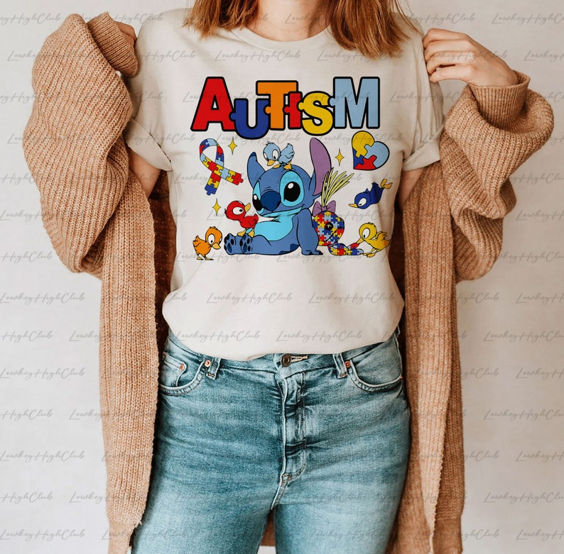 Op de afbeelding: Een wit T-shirt met het woord "AUTISME" in regenboogkleuren en een blauw Stitch-personage uit Lilo & Stitch. Het Stitch-personage wordt omringd door kleine vogels en een puzzelhart.