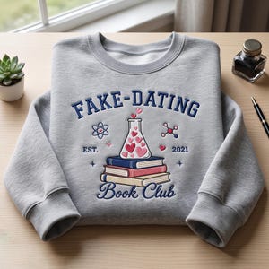 May include: A light grey crewneck sweatshirt featuring an embroidered design with the text FAKE-DATING BOOK CLUB, EST. 2021. The graphic includes a stack of three books topped with a laboratory flask filled with pink and red hearts, an atom, and a molecule.