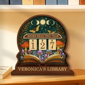 May include: A wooden book counter with a whimsical design. The counter displays "BOOKS READ THIS YEAR" above the numbers 127. The design includes mushrooms, butterflies, and the text "VERONICA'S LIBRARY".
