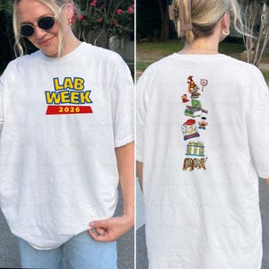 May include: White t-shirt with the text "LAB WEEK 2026" in blue, yellow, and red. The back of the shirt features a vertical design with various cartoon lab-related images. The shirt is a casual, oversized fit.
