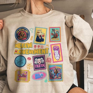 May include: A beige crewneck sweatshirt featuring a graphic print of various colorful postage stamp-style illustrations. The text "NEEEW ACHIEVEMENT" is printed in large yellow letters, surrounded by cartoon images of cats, dinosaurs, and abstract shapes.