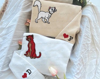 Könnte beinhalten: Zwei weiße Sweatshirts mit gestickten Katzenbildern. Ein Sweatshirt hat eine weiße Katze mit einem roten Herzen und dem Buchstaben "F" gestickt. Das andere Sweatshirt hat eine rote Katze mit einem roten Herzen und dem Buchstaben "D" gestickt.