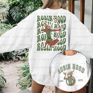 May include: White sweatshirt with a green and brown cartoon fox character holding a bow and arrow. The text "ROBIN HOOD" is repeated in a retro font on the back of the sweatshirt. The front of the sweatshirt has a circular design with the text "ROBIN HOOD EST. 1973" and a cartoon fox character.