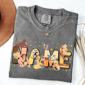 May include: Gray Comfort Colors t-shirt with a custom name design featuring cartoon characters. The word "NAME" is in yellow block letters with jungle-themed accents and character illustrations. The shirt has a crew neck and short sleeves.