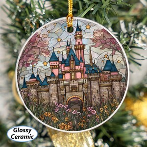 May include: A round, glossy ceramic ornament featuring a colorful stained-glass design of a fairytale castle with pink, blue, and gray tones. The castle is surrounded by flowers and stars, with a gold hanging cord.