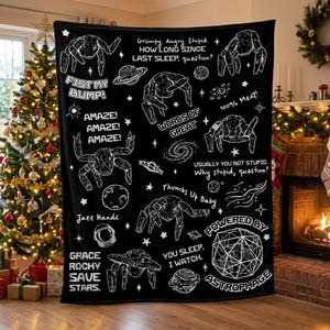 May include: A black throw blanket featuring white line art of space-themed illustrations, including planets, stars, and spider-like creatures, with text such as "Fist my bump!", "Amaze!", "100% Meat!", "Thumbs Up Baby", and "Powered by Astrophage".