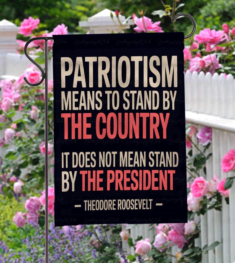 Puede incluir: Una bandera de jard&iacute;n negra con texto blanco y rojo que dice: "PATRIOTISMO SIGNIFICA APOYAR AL PA&Iacute;S, NO SIGNIFICA APOYAR AL PRESIDENTE - THEODORE ROOSEVELT -". La bandera est&aacute; colgada en un jard&iacute;n con flores rosas.
