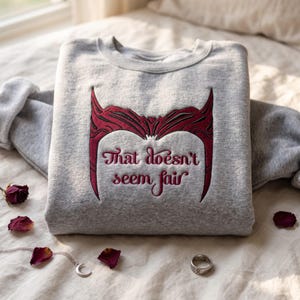 Op de afbeelding: Een grijs gemêleerd sweatshirt met ronde hals en een donkerrode geborduurde afbeelding van een puntige hoofdtooi, inclusief de tekst "That doesn't seem fair" in een cursief lettertype. Het ontwerp staat centraal op de borst van de trui.