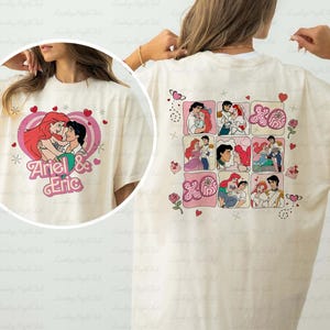 May include: Cream-colored t-shirt featuring a cartoon graphic of Ariel and Eric in a heart-shaped design. The back of the shirt displays a grid of illustrations with hearts and roses. The shirt is a casual wear item.