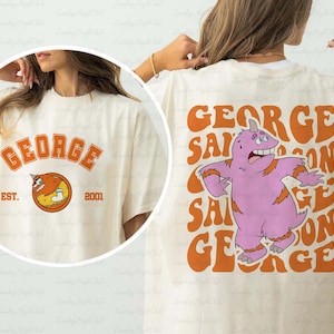 Könnte beinhalten: Cremefarbenes T-Shirt mit dem Namen "GEORGE" in orangefarbenen Buchstaben. Auf der Vorderseite befindet sich eine Cartoonfigur und der Text "EST. 2001". Auf der Rückseite steht "GEORGE SAMSON" mit einem lila Monster.