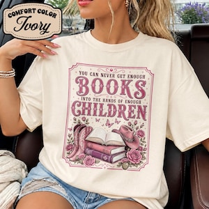 Könnte beinhalten: Ein elfenbeinfarbenes T-Shirt mit einem Grafikdruck und dem Text "YOU CAN NEVER GET ENOUGH BOOKS INTO THE HANDS OF ENOUGH CHILDREN." Das Design zeigt rosa Cowboystiefel, einen Stapel Bücher, einen rosa Cowboyhut, Rosen und Schmetterlinge.