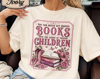 May include: An ivory t-shirt featuring a graphic print with the text "YOU CAN NEVER GET ENOUGH BOOKS INTO THE HANDS OF ENOUGH CHILDREN." The design includes pink cowboy boots, a stack of books, a pink cowboy hat, roses, and butterflies.