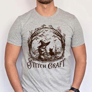 May include: A heather grey short-sleeve t-shirt featuring a dark brown graphic print of a witch knitting with a black cat. The design includes a crescent moon, stars, bats, and trees, with the text STITCH CRAFT printed in a stylised font below.